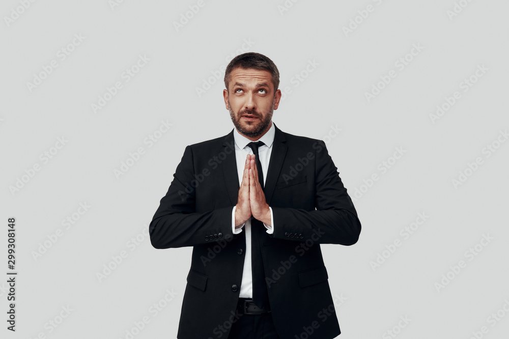 Charming young man in full suit keeping hands clasped while standing against grey background Charming young man in full suit keeping hands clasped while standing against grey background