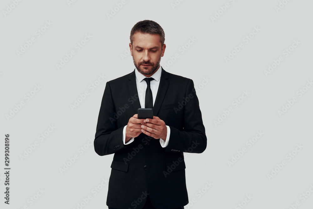 Charming young man in full suit using smart phone while standing against grey background Charming young man in full suit using smart phone while standing against grey background
