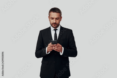Charming young man in full suit using smart phone while standing against grey background