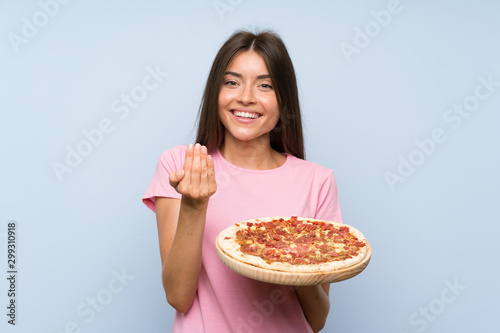 Pretty young girl holding a pizza over isolated blue wall inviting to come with hand. Happy that you came