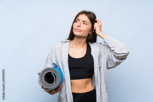 Fototapeta Naklejka Na Ścianę i Meble -  Young sport girl with mat over isolated blue background having doubts and with confuse face expression
