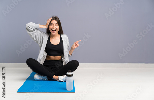 Young sport woman sitting on the floor with mat surprised and pointing finger to the side