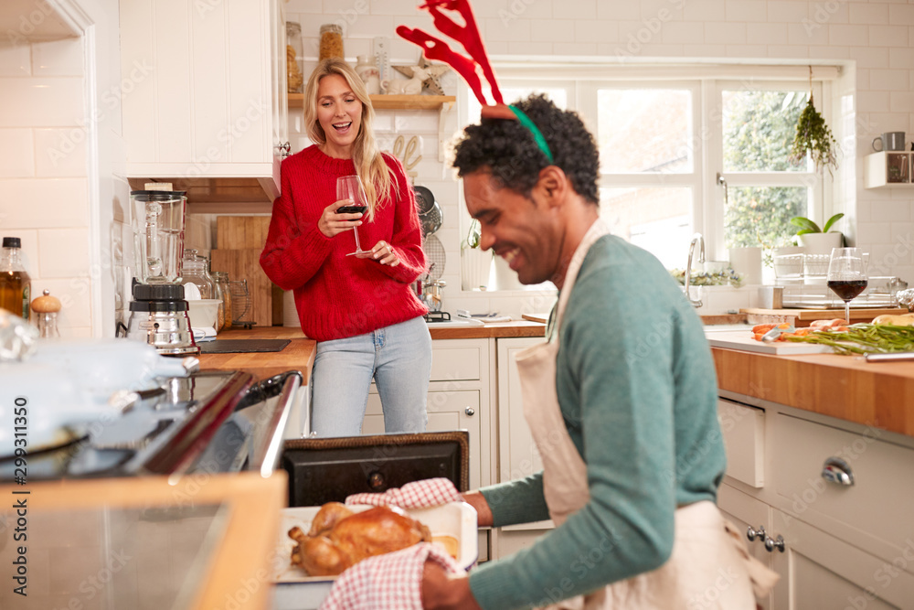 Man Taking Turkey Out Of Oven As Couple Drink Wine And Prepare Dinner On Christmas Day Man Taking Turkey Out Of Oven As Couple Drink Wine And Prepare Dinner On Christmas Day