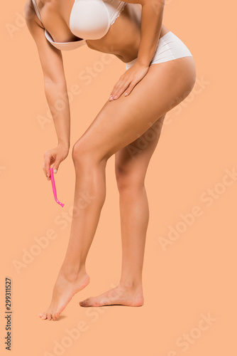 cropped view of woman shaving legs while standing isolated on beige