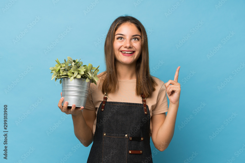 Young brunette girl over isolated blue background taking a flowerpot and pointing up Young brunette girl over isolated blue background taking a flowerpot and pointing up