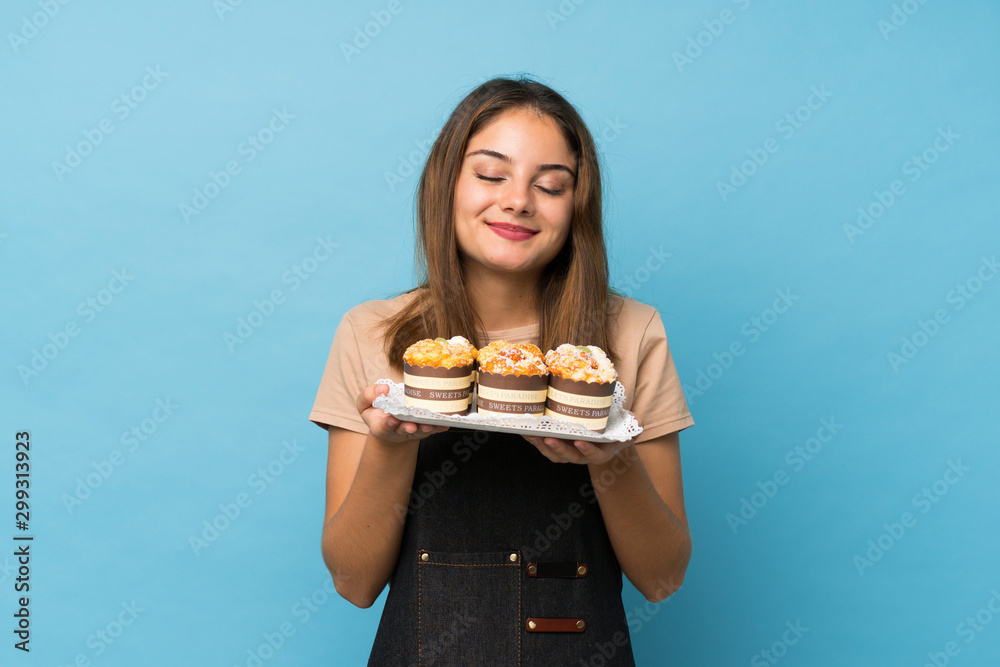 Young brunette girl over isolated blue background holding mini cakes enjoying the smell of them Young brunette girl over isolated blue background holding mini cakes enjoying the smell of them