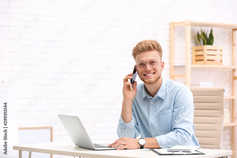 Handsome young man talking on phone while working with laptop at table in office Handsome young man talking on phone while working with laptop at table in office