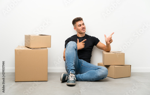 Handsome young man moving in new home among boxes pointing finger to the side