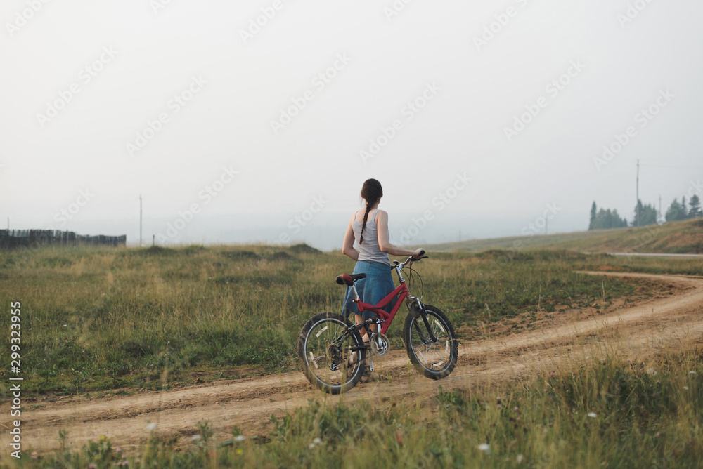 white caucasian young woman in casual clothing walking with bicycle on village road, view from back in full body size, lifestyles stock photo image