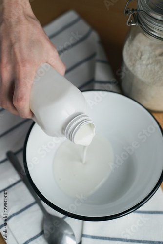 caucasian womans hand holds plastic bottle, pours kefir in white metal bowl on wooden table, cooking homemade oladyi, pancakes with wholegrain flour, top view of vertical life style stock photo image