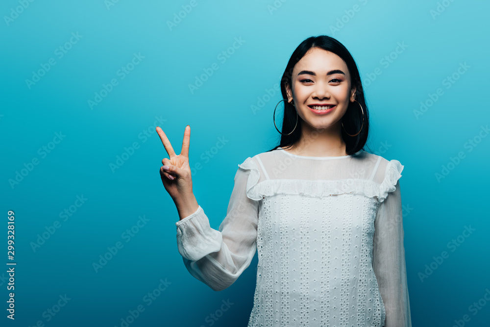 happy brunette asian woman showing peace sign on blue background happy brunette asian woman showing peace sign on blue background