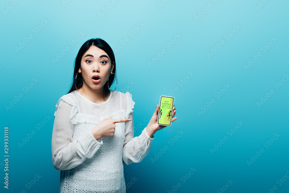 shocked asian woman pointing with finger at smartphone with fashion shopping app on blue background shocked asian woman pointing with finger at smartphone with fashion shopping app on blue background