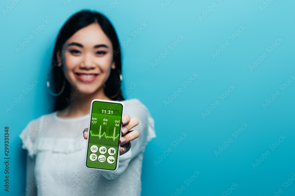 selective focus of smiling asian woman in white blouse holding smartphone with healthcare app on blue background selective focus of smiling asian woman in white blouse holding smartphone with healthcare app on blue background