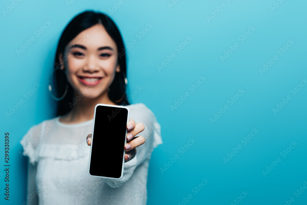 selective focus of smiling asian woman in white blouse holding smartphone with blank screen on blue background selective focus of smiling asian woman in white blouse holding smartphone with blank screen on blue background