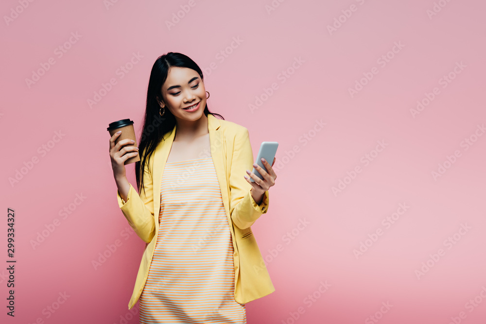 smiling asian woman in yellow outfit holding paper cup and using smartphone isolated on pink