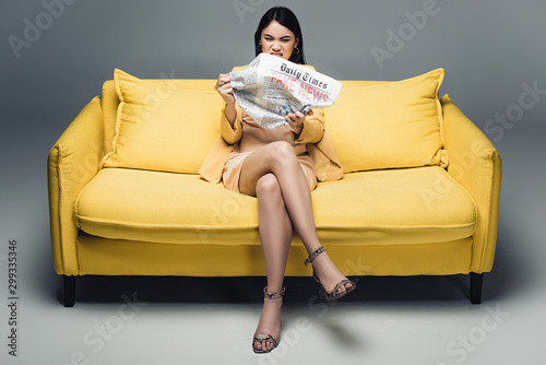 aggressive asian businesswoman sitting on yellow sofa and reading newspaper with fake news on grey background