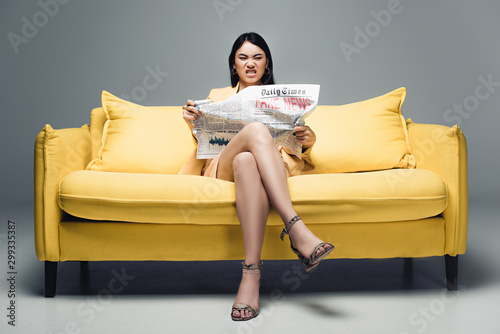 angry asian businesswoman sitting on yellow sofa and reading newspaper with fake news on grey background