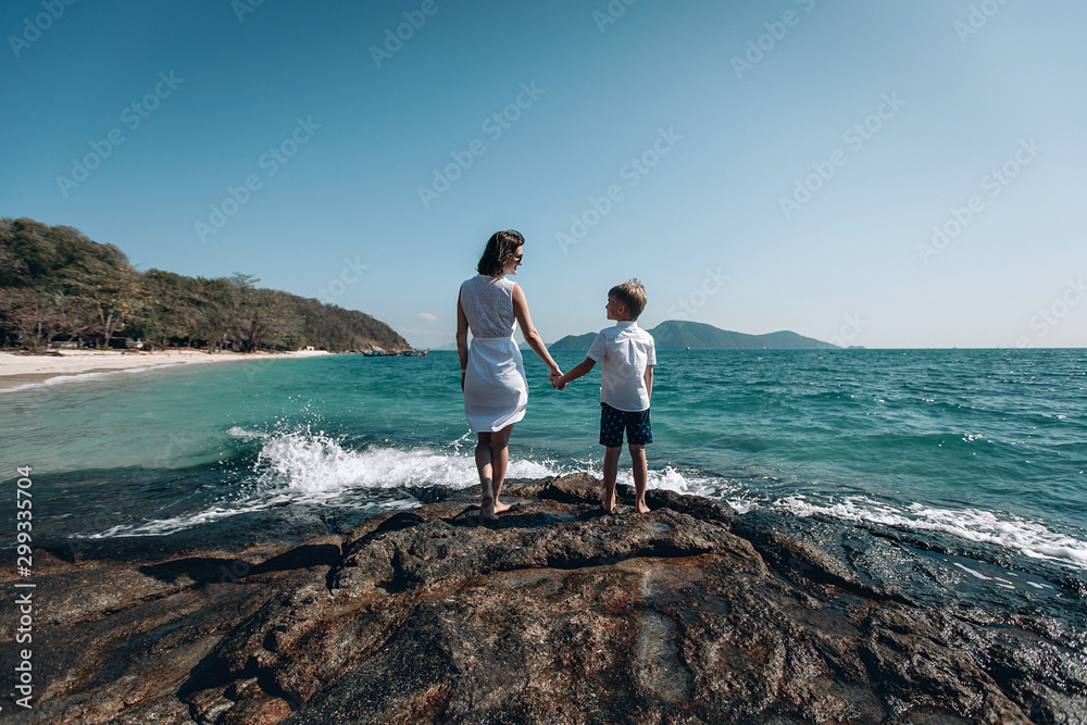 Loving mother and her little son hold hands and and look at the turquois sea standing on rocky beach