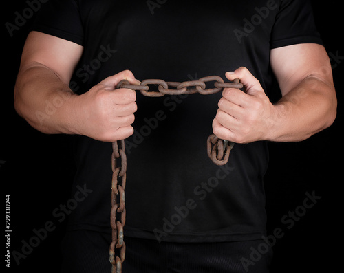 adult man in black clothes stands upright with strained muscles and holds a rusty metal chain