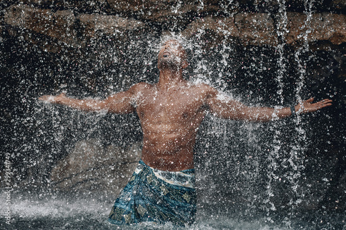 Fit and handsome topless Arabic man under the waterfall  huge rocks and water  splashes and flecks  natural beauty concept.