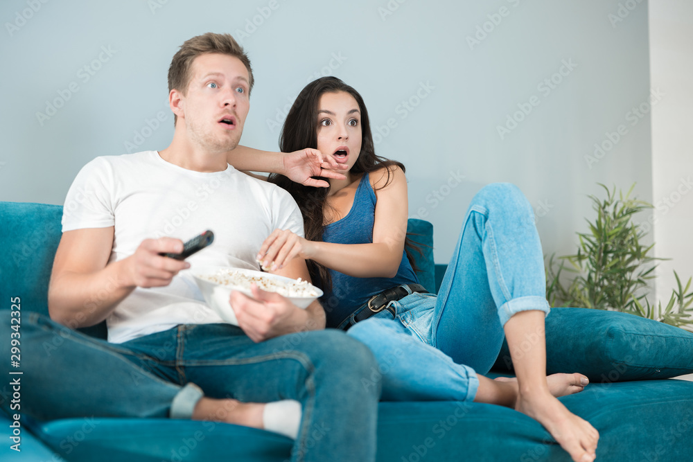 young couple beautiful brunette woman and handsome man watching scary movie on the sofa eating pop corn looking frightened cozy lifestyle