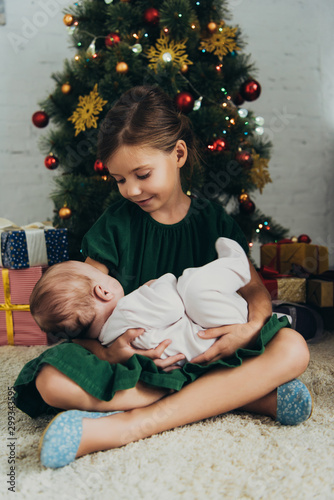 adorable child holding cute little sisiter while sitting near Christmas tree and gift boxes photo