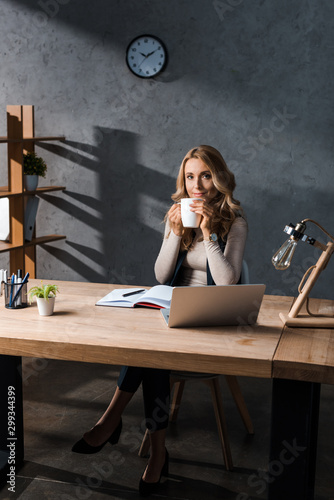 attractive and blonde businesswoman sitting at table and holding cup in office