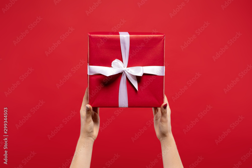 cropped view of woman holding wrapped Christmas present with white ribbon isolated on red background. Christmas concept