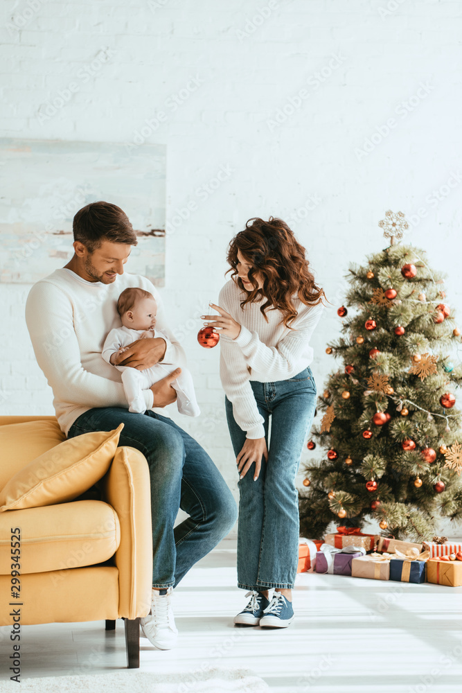 mother showing christmas ball to infant sitting on fathers hands near decorated christmas tree mother showing christmas ball to infant sitting on fathers hands near decorated christmas tree