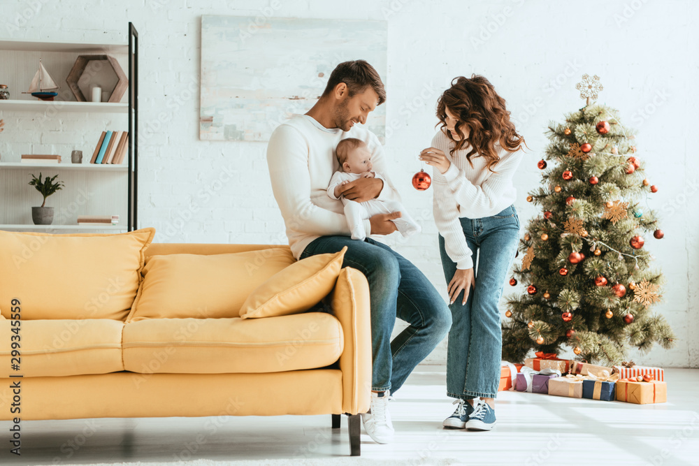 Obraz premium Woman showing Christmas ball to little child sitting on fathers hands near decorated Christmas tree