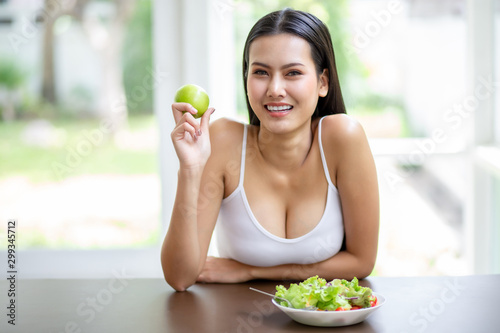 happy sexy young asian woman in white singlet smiling , holding green apple and eating fresh organic salad . A beautiful girl have breakfast healthy good food in morning at home. weight loss concept.