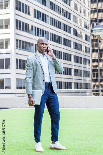 Young African American Businessman Street Fashion in New York City, wearing gray patterned blazer, white shirt, blue pants, sneakers, carrying laptop computer, standing on green lawn, talking on phone