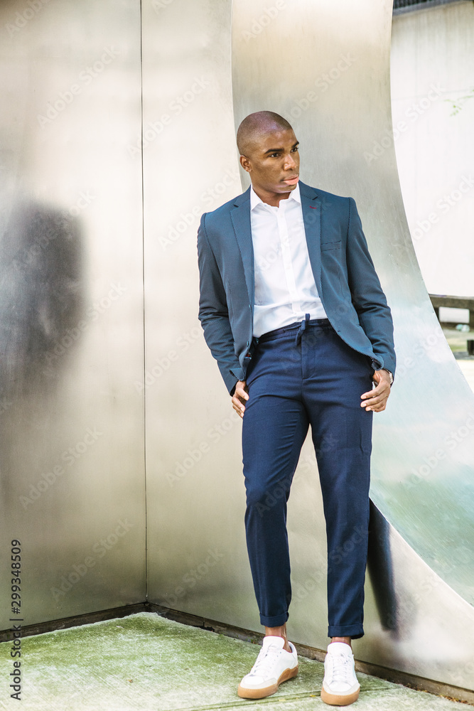 Portrait of Young African American Man in New York City. Wearing blue  blazer, white shirt, blue pants, white sneakers, young black college  student standing against silver metal wall on campus, think.. Stock