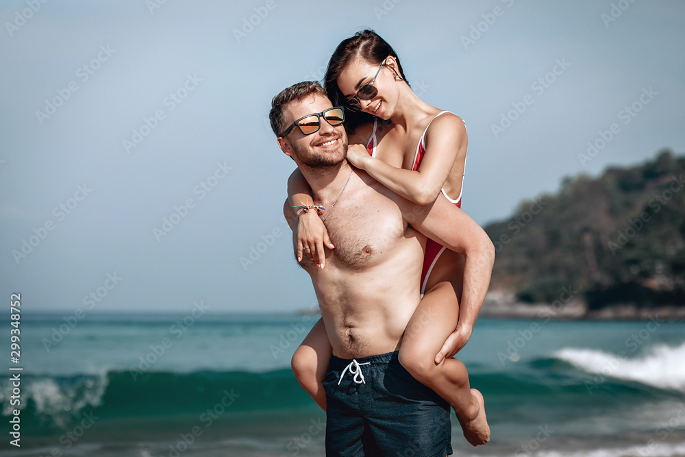 Funny couple on the beach, the muscular man holds his beloved girlfriend on his back, both in swimwear, smiling and tanned; honeymoon concept. Funny couple on the beach, the muscular man holds his beloved girlfriend on his back, both in swimwear, smiling and tanned; honeymoon concept.