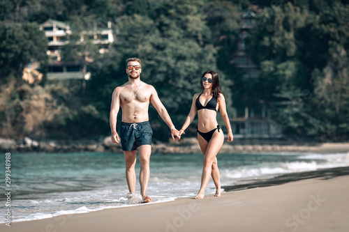 Loving couple on honeymoon walks along the sunny exotic beach holding hands  sunglasses and swimwear on  romance concept.