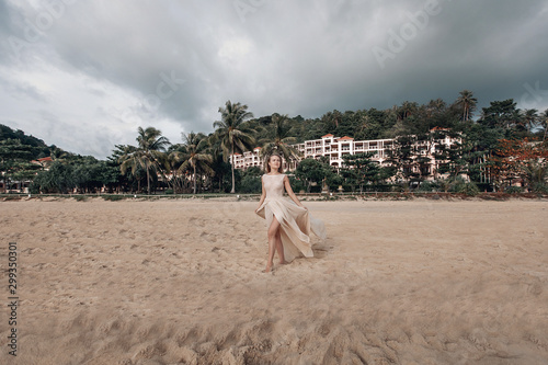 Bilde på lerret Elegant young lady in beige light dress standing on the sandy beach barefoot, costal hotel behind her; fashion concept