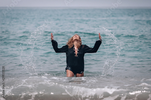 Happy smiling woman with thick blond wavy hair  wearing black long shirt stands in the sea near the shore playing with water  splashes and splatter.