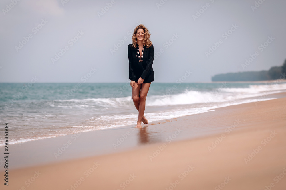 Beautiful slim woman with thick wavy hair, wearing black long shirt walks along the beach ankle-deep in the water.