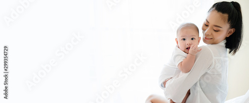 Portrait of beautiful Asian mother hugging his newborn son in the bedroom. Baby is adorable. Mom is smiling happily with the child healthy. Nursery for young children. Banner with copy space
