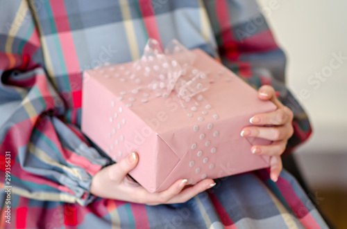 The hands of a girl in a checkered dress are holding an elegant, delicate pink gift with an airy, transparent polka dot ribbon. New year greeting card. Christmas package from Santa Claus. Copy space