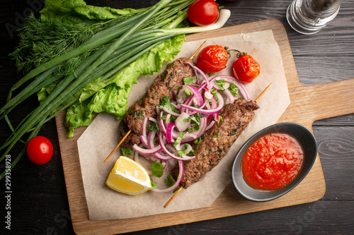 lula kebab of beef on skewers with onions, greens, ketchup and lemon on a board
