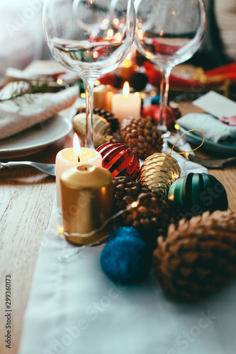 Table served for Christmas dinner in living room.  Close up view  table setting plates  branch decoration  candles and gliterring toys on wooden table background. Winter decoration