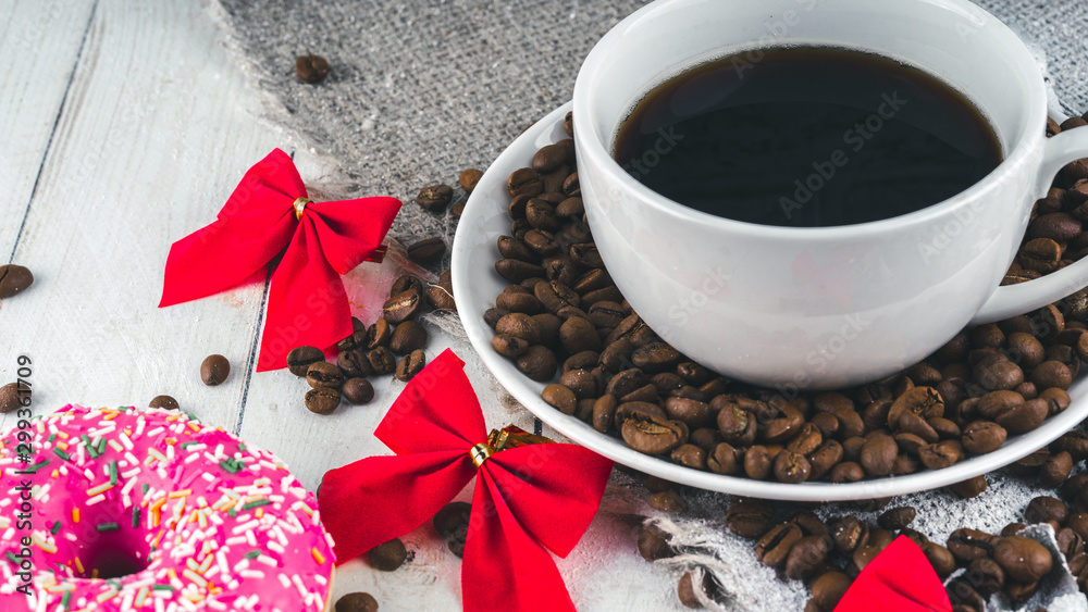 A hot cup of coffee with a doughnut and the coffee beans on the table. Composition of a delicious coffee and sweet snack. A hot cup of coffee with a doughnut and the coffee beans on the table. Composition of a delicious coffee and sweet snack.
