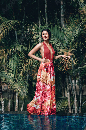 Attractive Asian girl in red dress posing near the infinity pool; recreation concept.