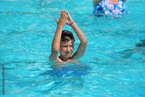 Smiling Caucasian boy having fun in swimming pool at resort. He dancing aqua Zumba and claping hands.