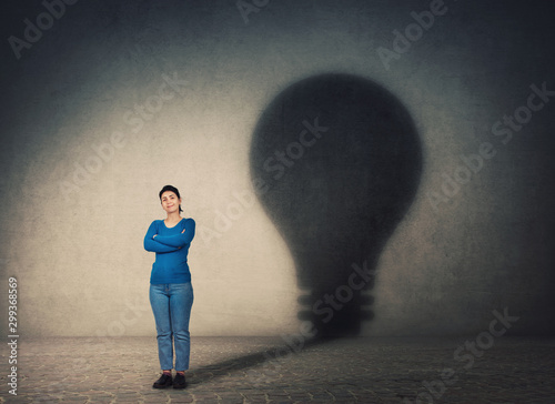 Confident woman, keeps arms crossed, casting a lightbulb shape shadow on the wall. Ambition and business idea concept. Genius innovation, motivation and inner power symbol. photo