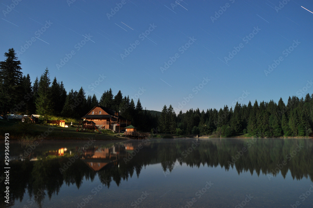 Fototapeta premium Forest and cottage reflecting in the lake at night