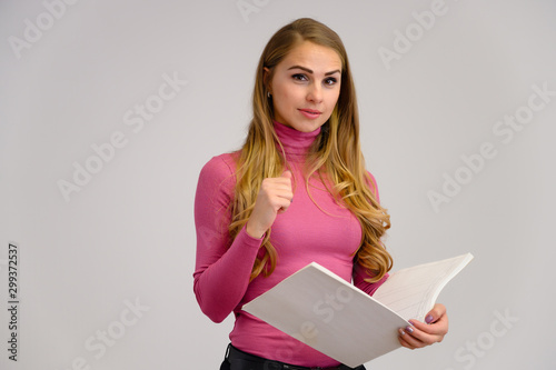 Close-up portrait of a pretty blonde girl with long curly hair standing in the studio on a white background with emotions in different poses in a pink sweater with a folder in her hands.