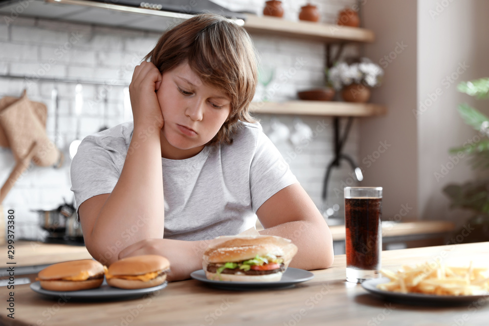 Emotional overweight boy at table with fast food in kitchen
