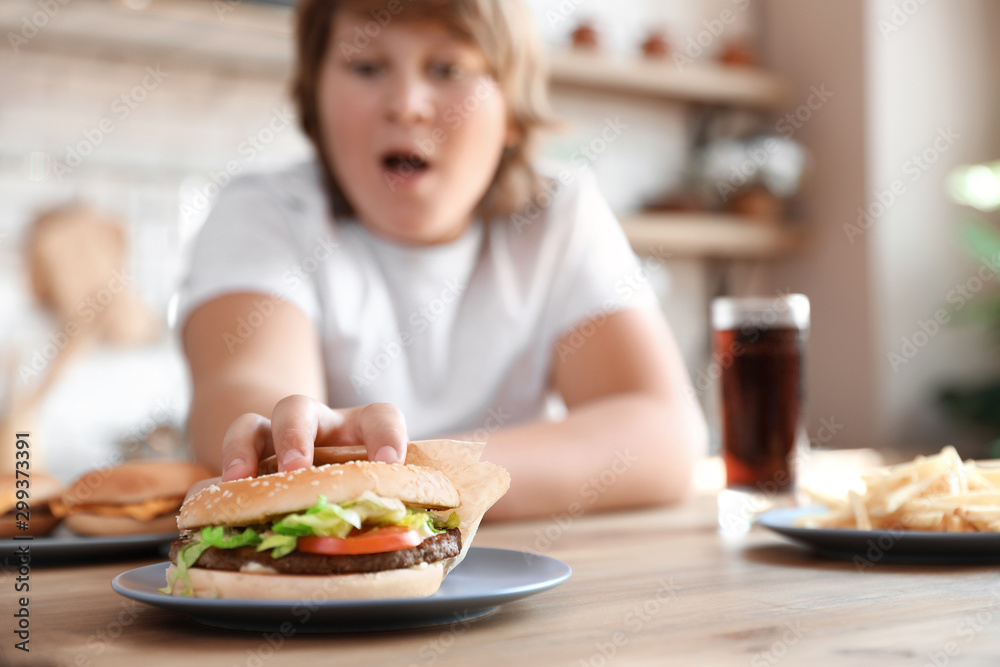 Emotional overweight boy reaching for burger at table in kitchen Emotional overweight boy reaching for burger at table in kitchen